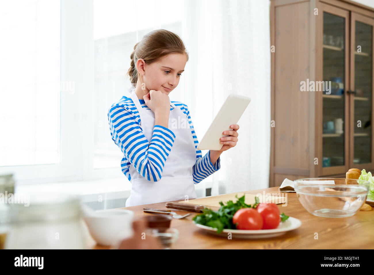Curious little girl wrapped up in reading recipe with help of digital tablet while standing at wooden kitchen table in order to make surprise for Moth Stock Photo