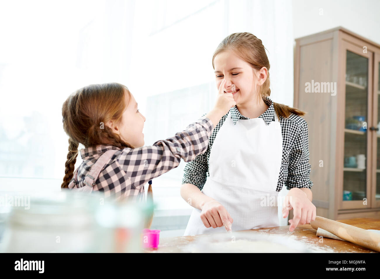 Profile view of cute little girl smearing face of her elder sister with flour while preparing pie together, interior of spacious kitchen on background Stock Photo