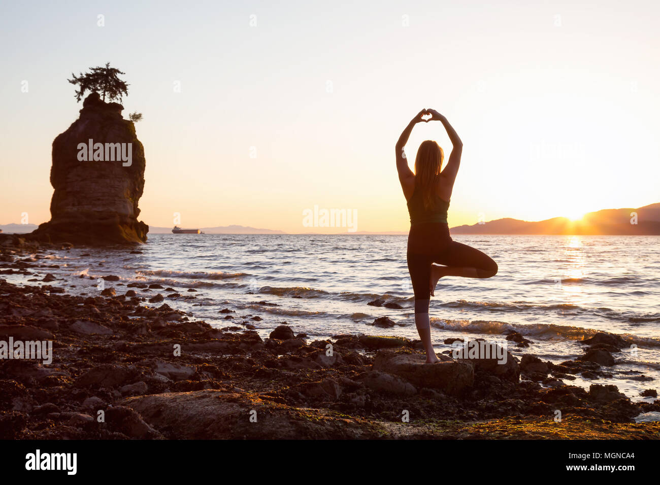 Young Woman practicing yoga on a rocky shore during a vibrant sunset. Taken in Stanley Park, Vancouver, British Columbia, Canada. Stock Photo