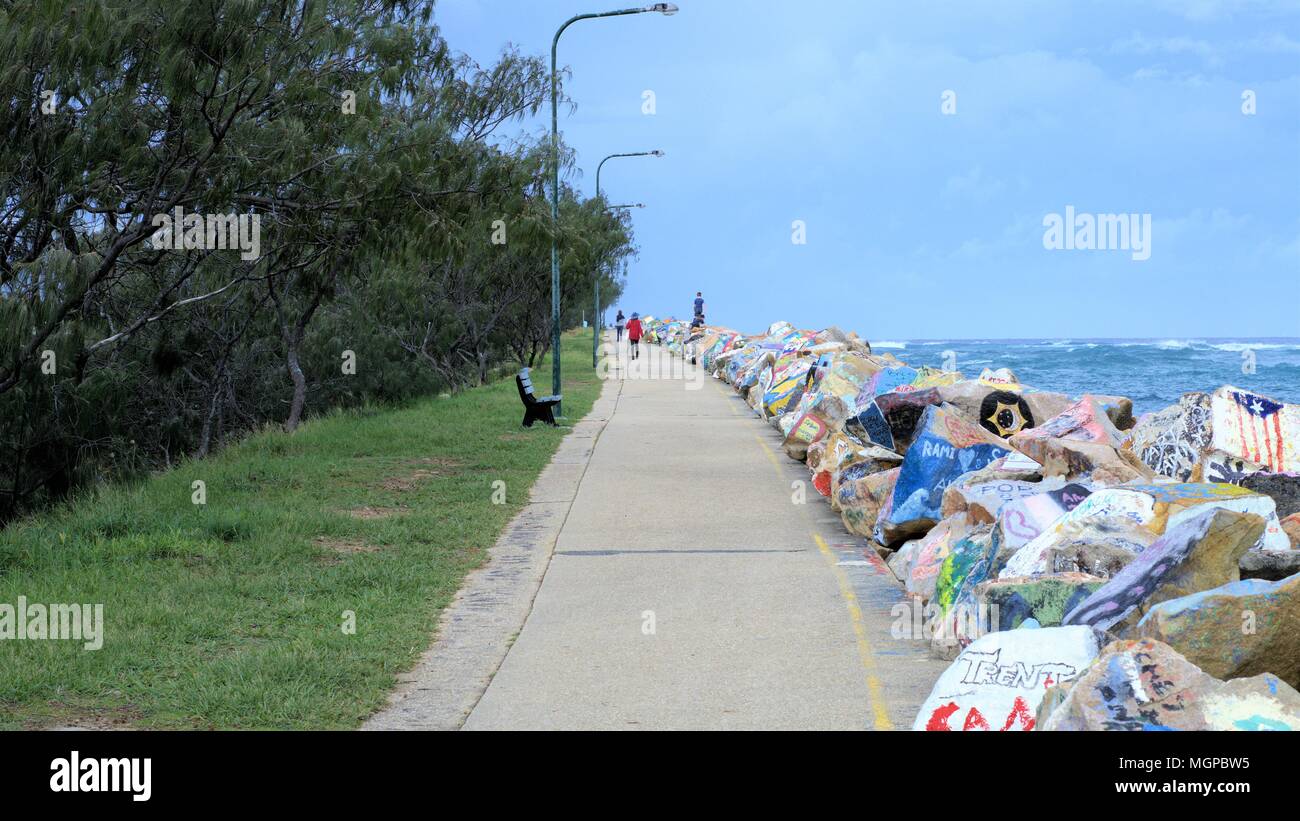 People walking on breakwater on river in Australia. Grey clouds rainy weather, concrete path, rocks, painted rocks, grass, trees Stock Photo