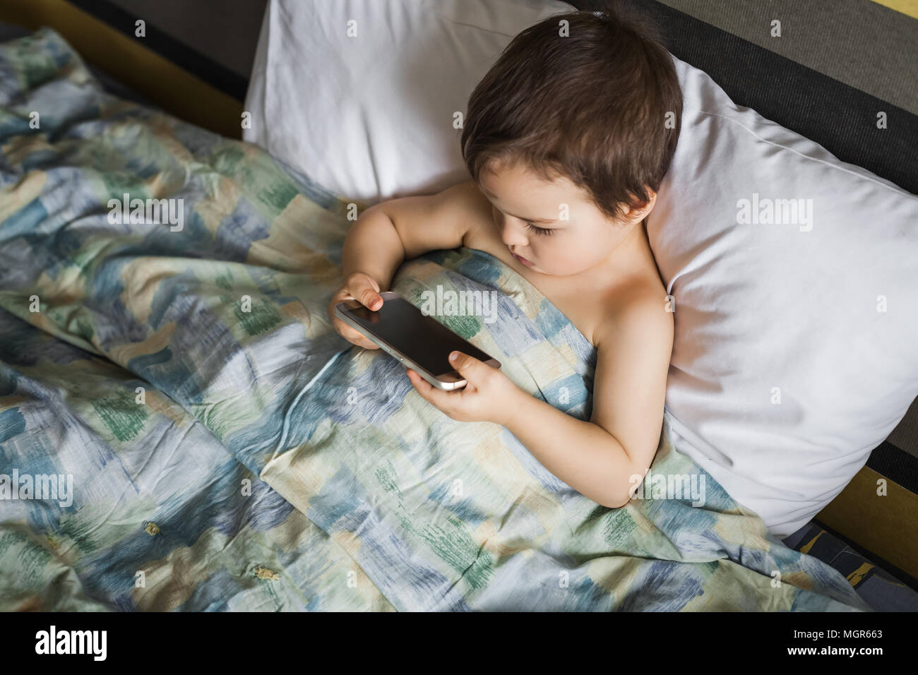 little smart boy holding playing mobile phone lying on bed in morning ...