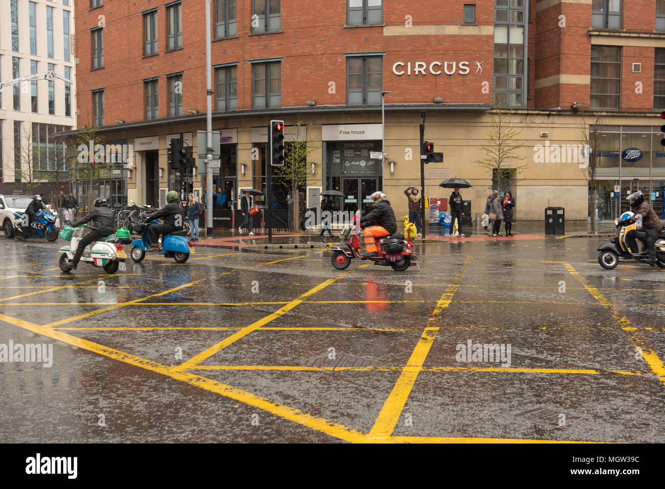 Group of scooter riders navigating a wet city intersection on a rainy day in front of Circus restaurant, Sheffield city centre, South Yorkshire, Engla Stock Photo