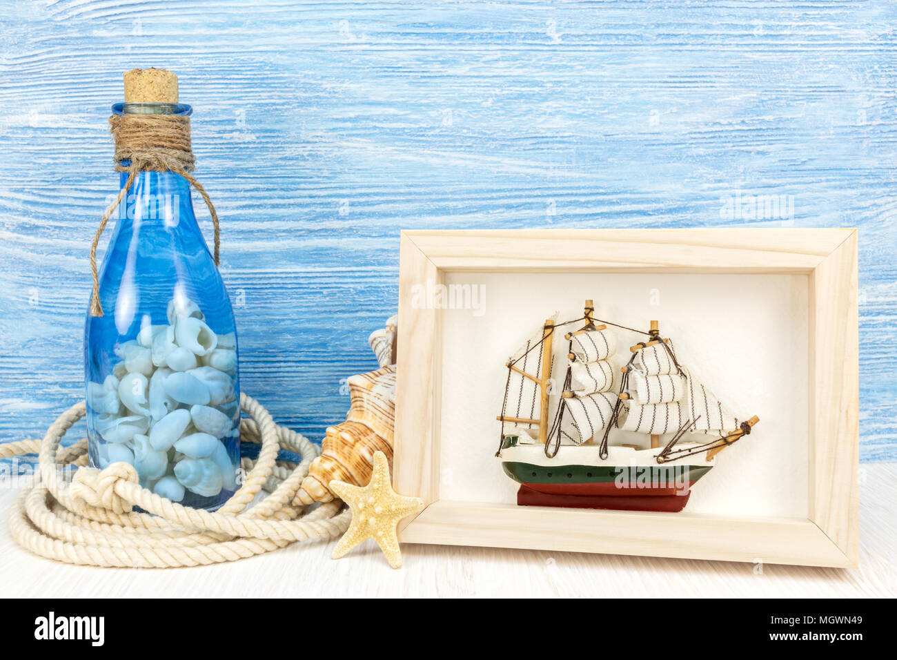 summer vacation concept – bottle with seashells, photo frame with sailboat, starfish and marine rope against blue wooden background Stock Photo