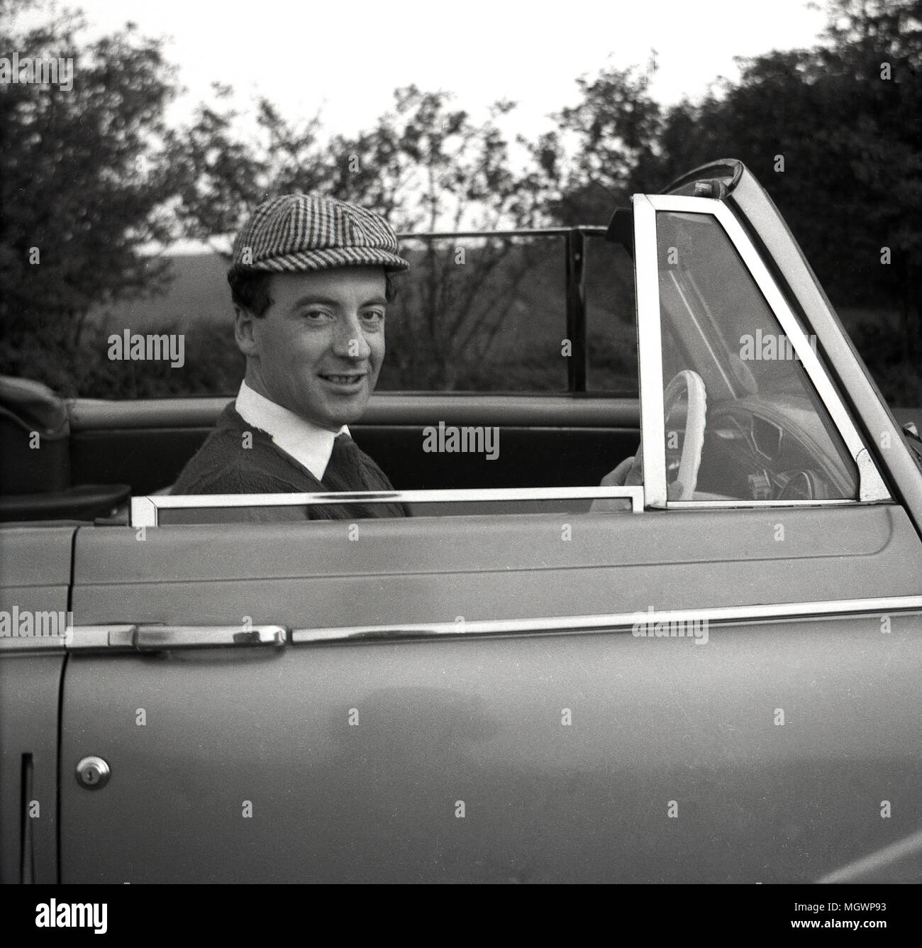 1955, historical, adult male driver sitting hands at the wheel of his soft-top motorcar wearing a fashionable 'sportsmans' version of the famous deerstalker cloth cap, made popular by the fiction detective, Sherlock Holmes, England, UK. Stock Photo