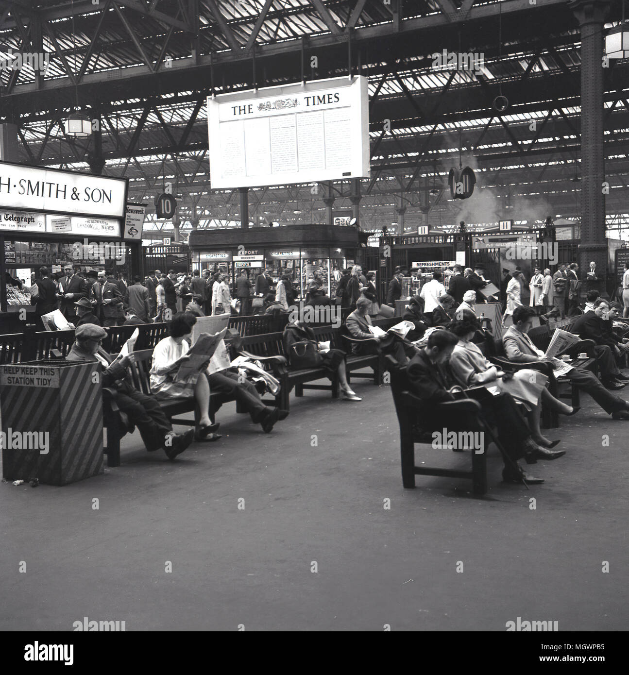 1950s, historical, rail passengers at St Pancras railway station sit on benches reading newspapers while awaiting their trains, London, England, UK. As can be seen in distance, it was very much still the era of steam, which can be seen arising from the platforms. The station was built by Midland Railway as their own central London terminus and opened in 1868, with its famous single-structure roof or trainshed designed by William Henry Barlow. Stock Photo