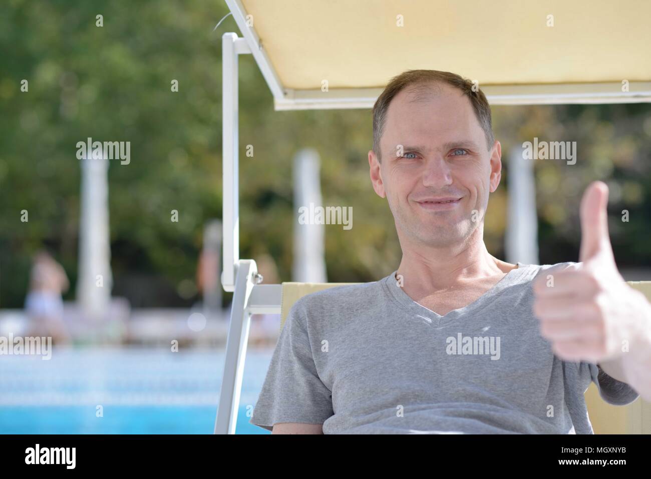 Man resting in a chaise lounge against a swimming pool and giving thumbs up sign Stock Photo