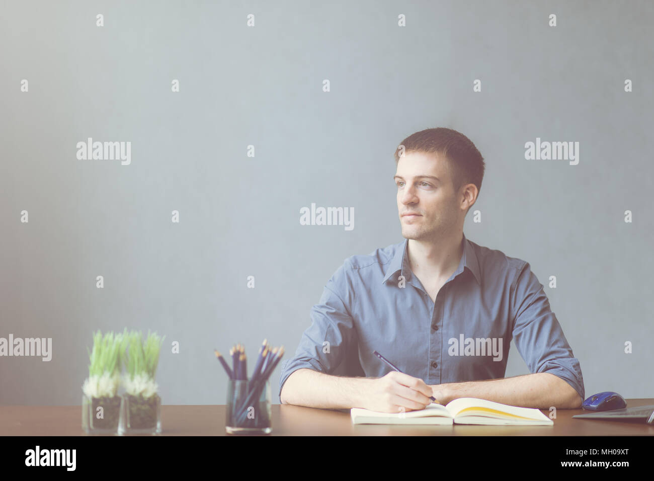 Young businessman caucasians sitting at desk office table and taking notes in notebook. Writing and looking out the window on the left and happy smile Stock Photo