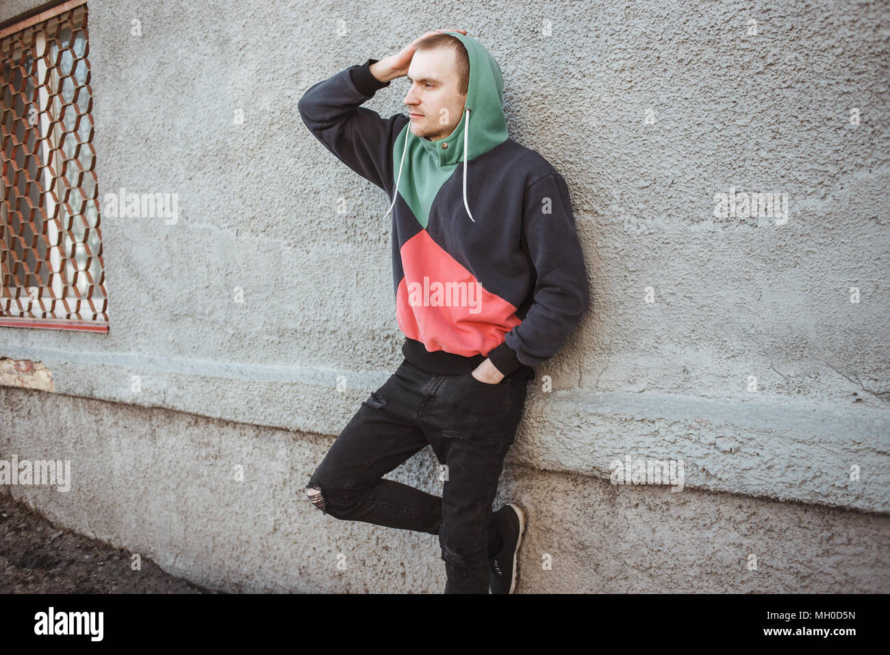Young man wearing a hoodie standing by grey wall and looking into the distance. Stock Photo