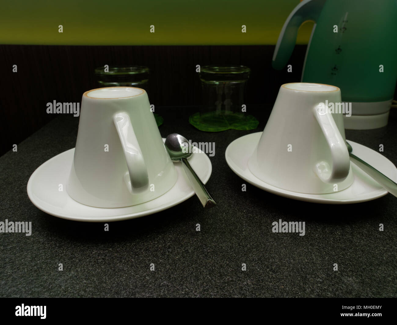 Two white coffee cups and tea spoons were put upside down over dark background with water glasses and kettle in the background with concepts of luxury, relaxation, wellness, and calm Stock Photo