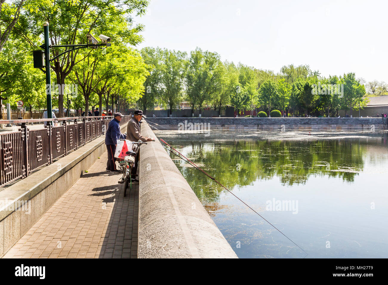 A Chinese man fishing with friends in the moat which circles the Forbidden City, Beijing China. Above them a surveillance camera keeps watch. Stock Photo