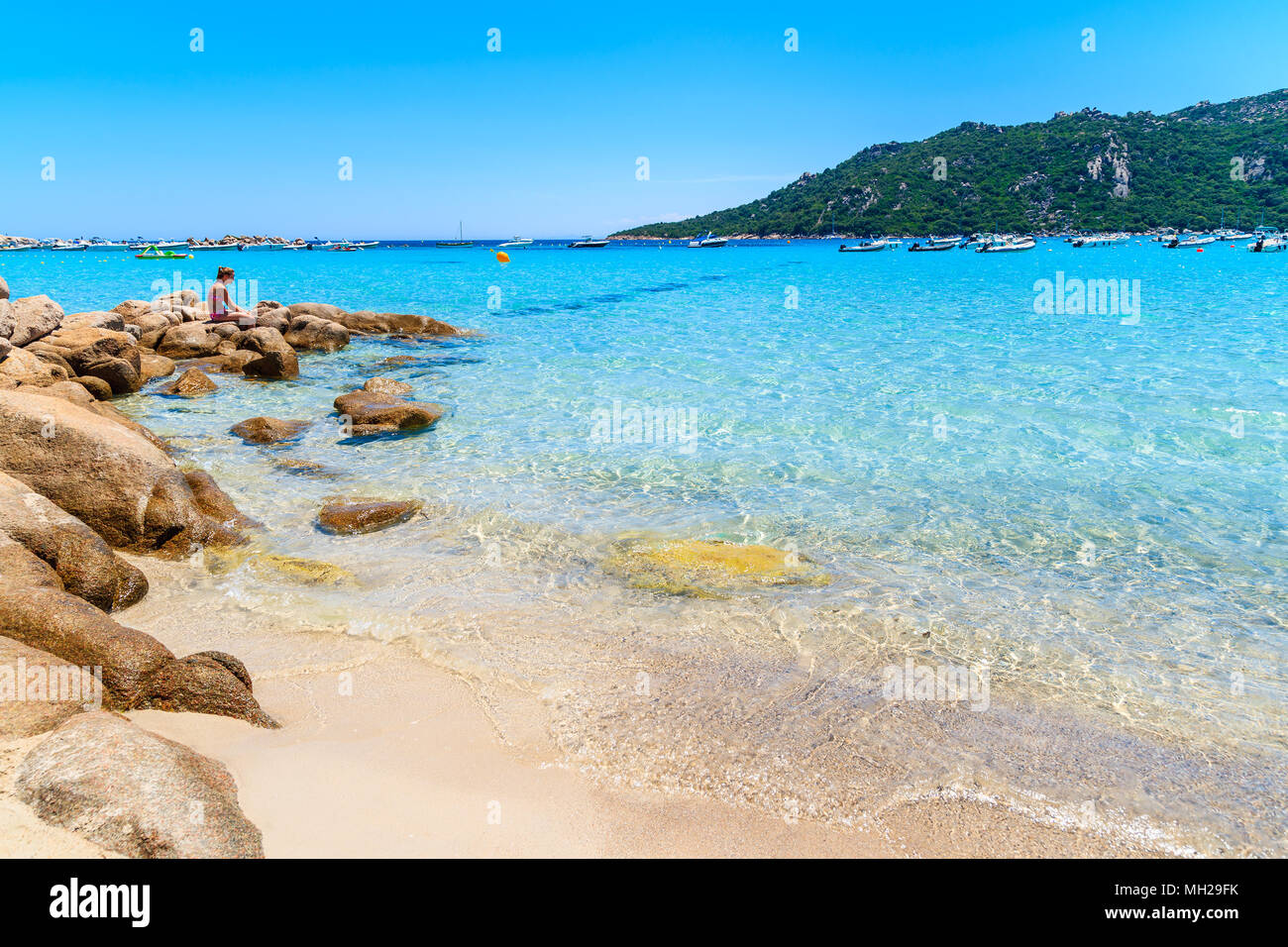 SANTA GIULIA BEACH, CORSICA ISLAND - JUN 22, 2015: Young woman sitting on rocks on beautiful beach with crystal clear water. This French island is pop Stock Photo