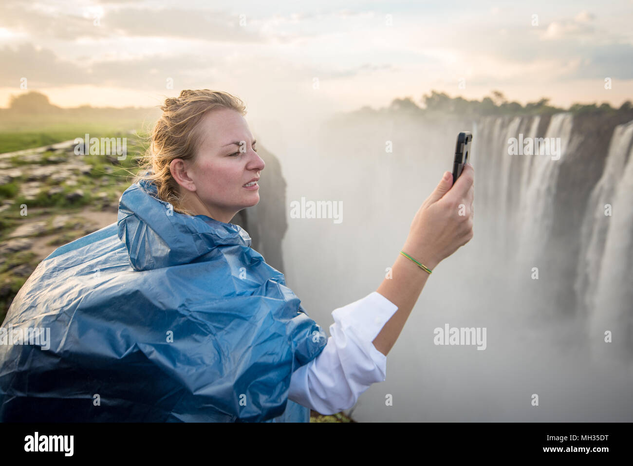 A young woman takes a photo in front of the rushing waters of Victoria Falls which spray mist into the air contributing to the areas thick, lush veget Stock Photo