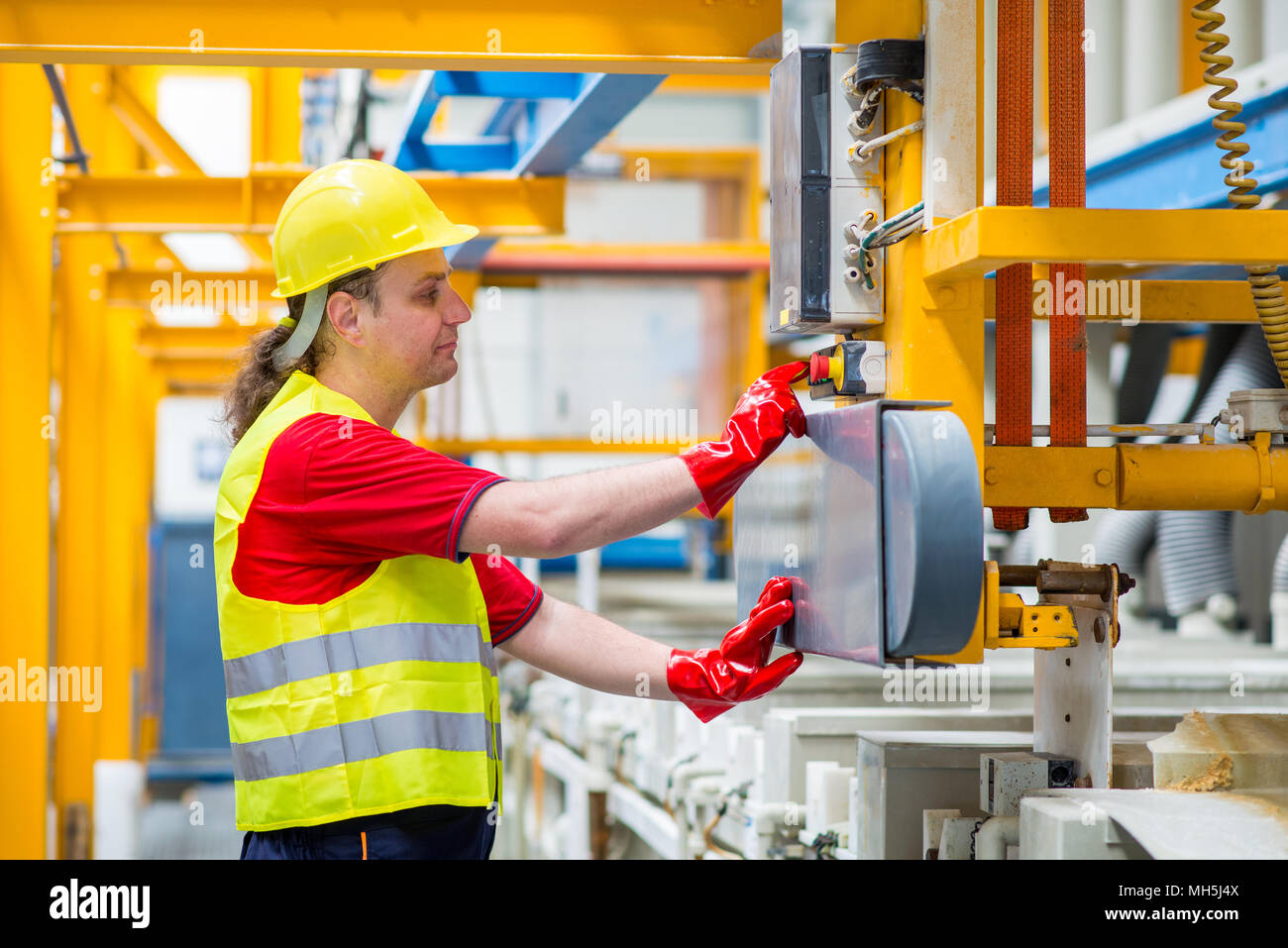 Worker in a factory pressing a red button. Worker wearing yellow reflective suit, yellow helmet and red working gloves Stock Photo