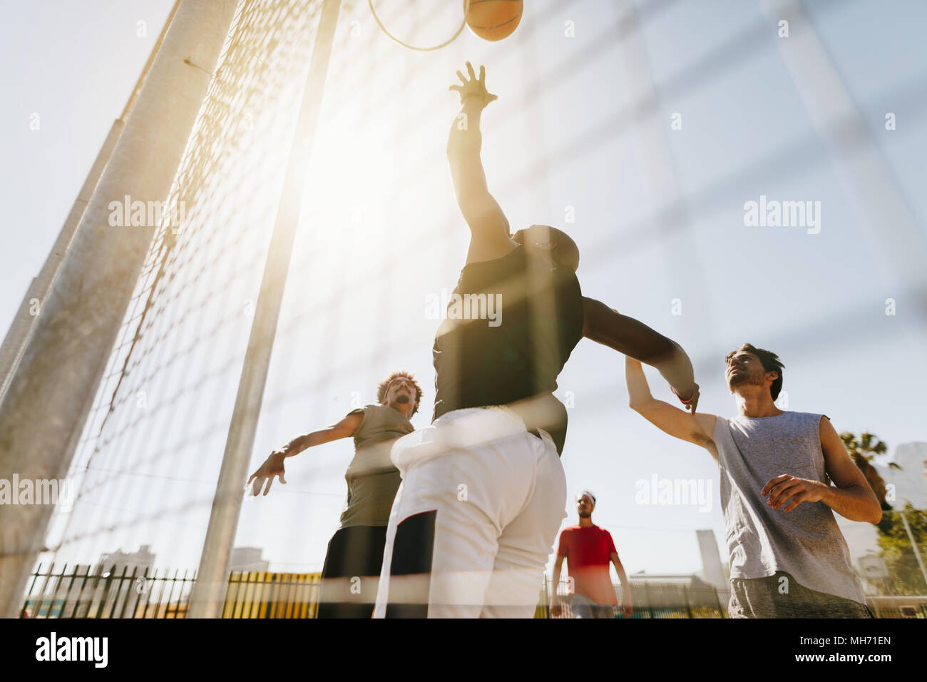 Four men playing basketball in a basketball court on a sunny day. Man jumping high to throw the ball in the basket. Stock Photo