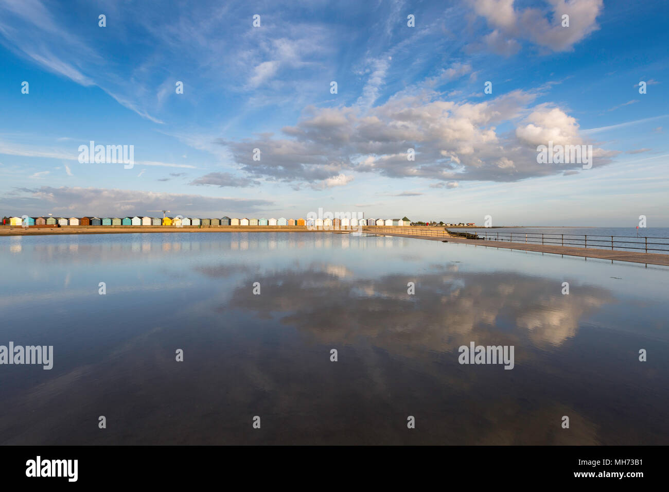 Dramatic clouds, the colourful beach huts and a blue sky reflected in the waters of the tidal pool, Brightlingsea, Essex, UK. Stock Photo