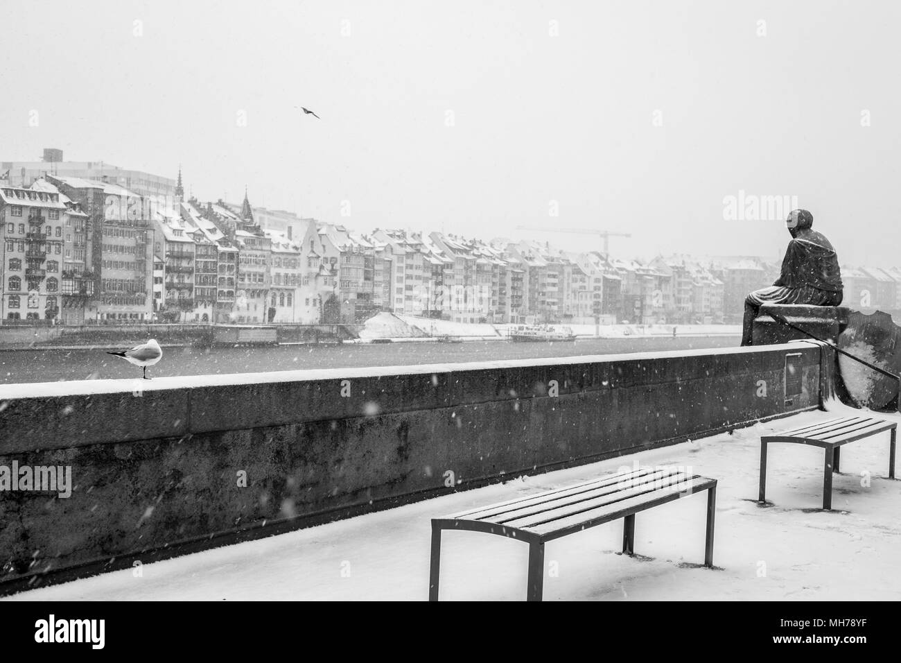 City Houses Near River In Cold, Snow Covered Winter Setting With Statue, Bench, Black and White Stock Photo