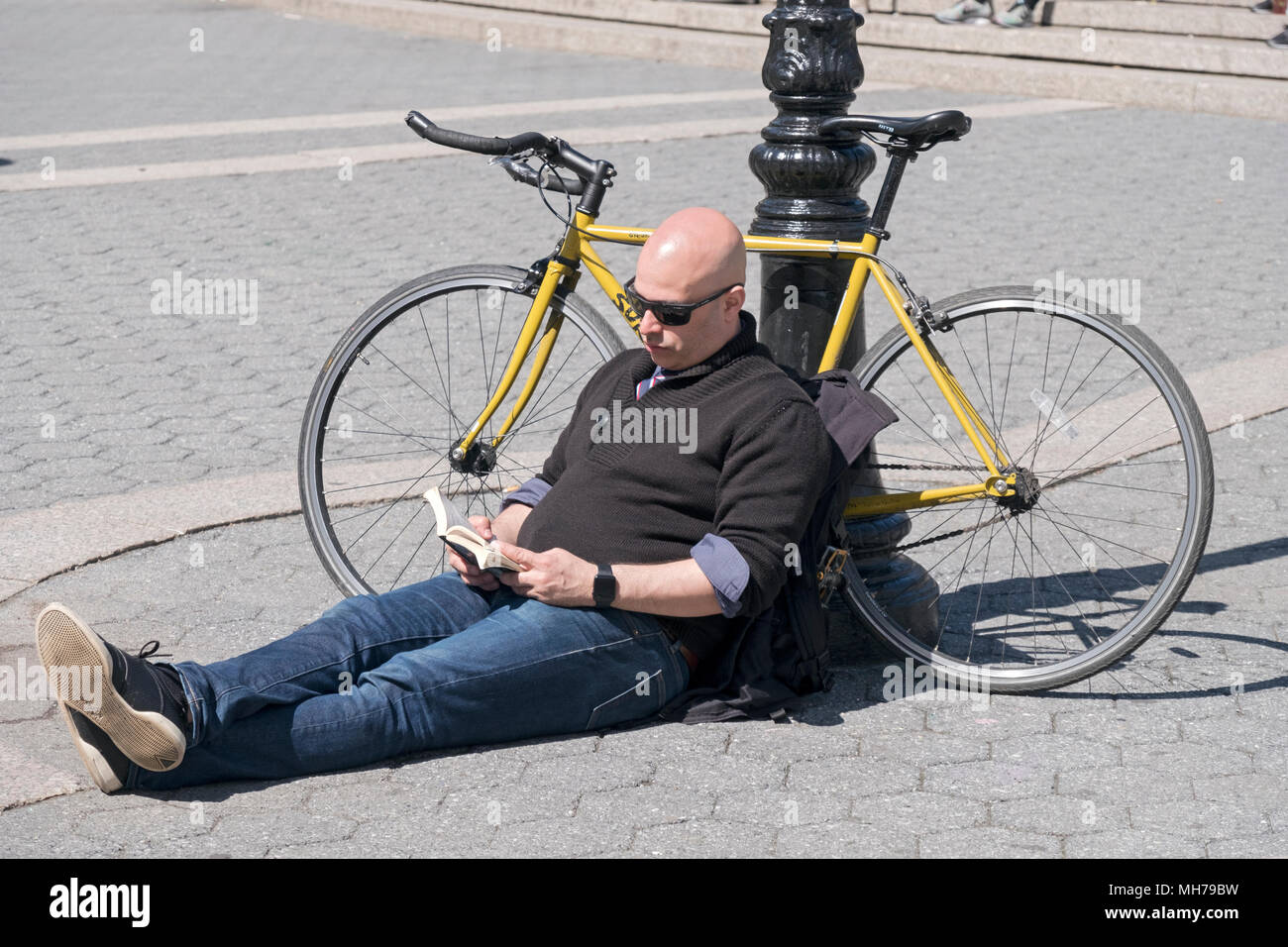 A man sits reading a book and leaning on his bicycle on a warm spring day. In Union Square Park in Manhattan, New York City. Stock Photo