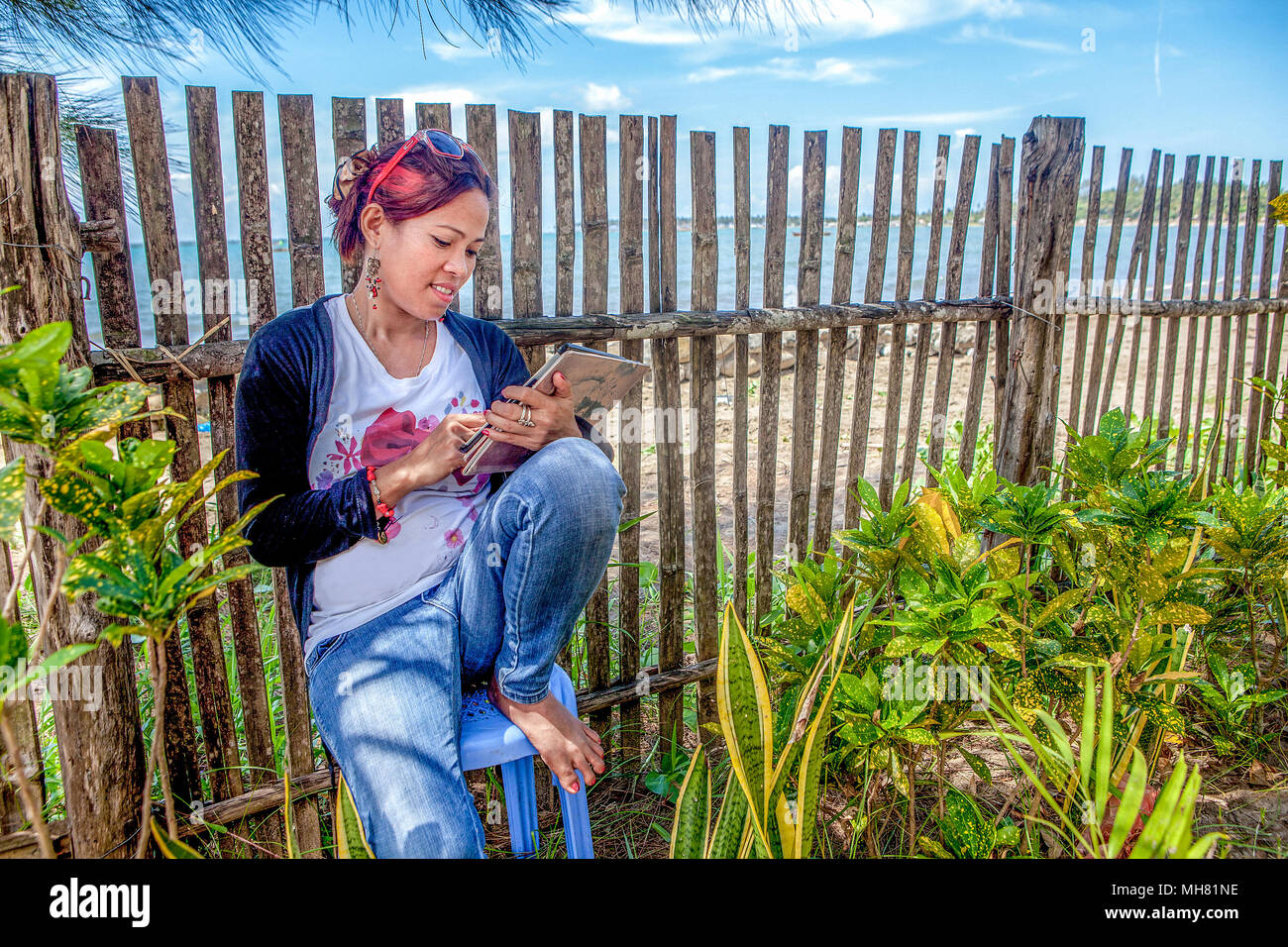 An Asian woman sits against a fence at a beach resort sending and receiving email on her iPad on Palawan Island, Philippines. Stock Photo