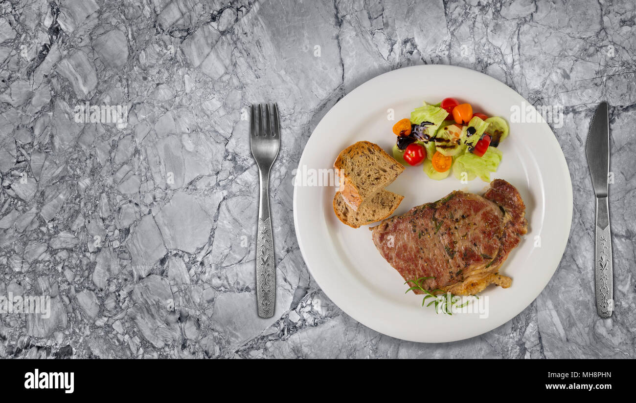 Ripened seasoned beef rump or striploin steak cooked on white plate with salad and home made bread. Top view on granite background with copy space. Stock Photo