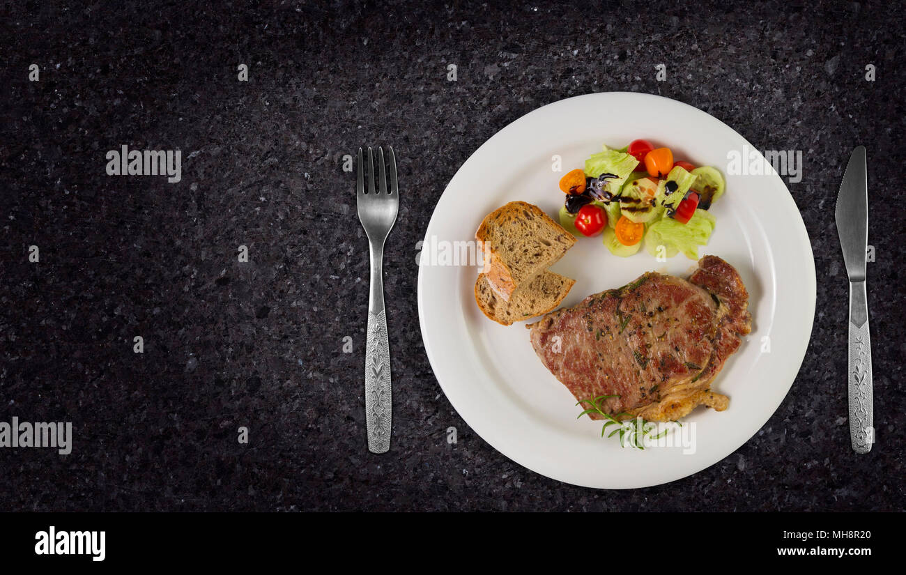 Ripened seasoned beef rump or striploin steak cooked on white plate with salad and home made bread. Top view on granite background with copy space. Stock Photo