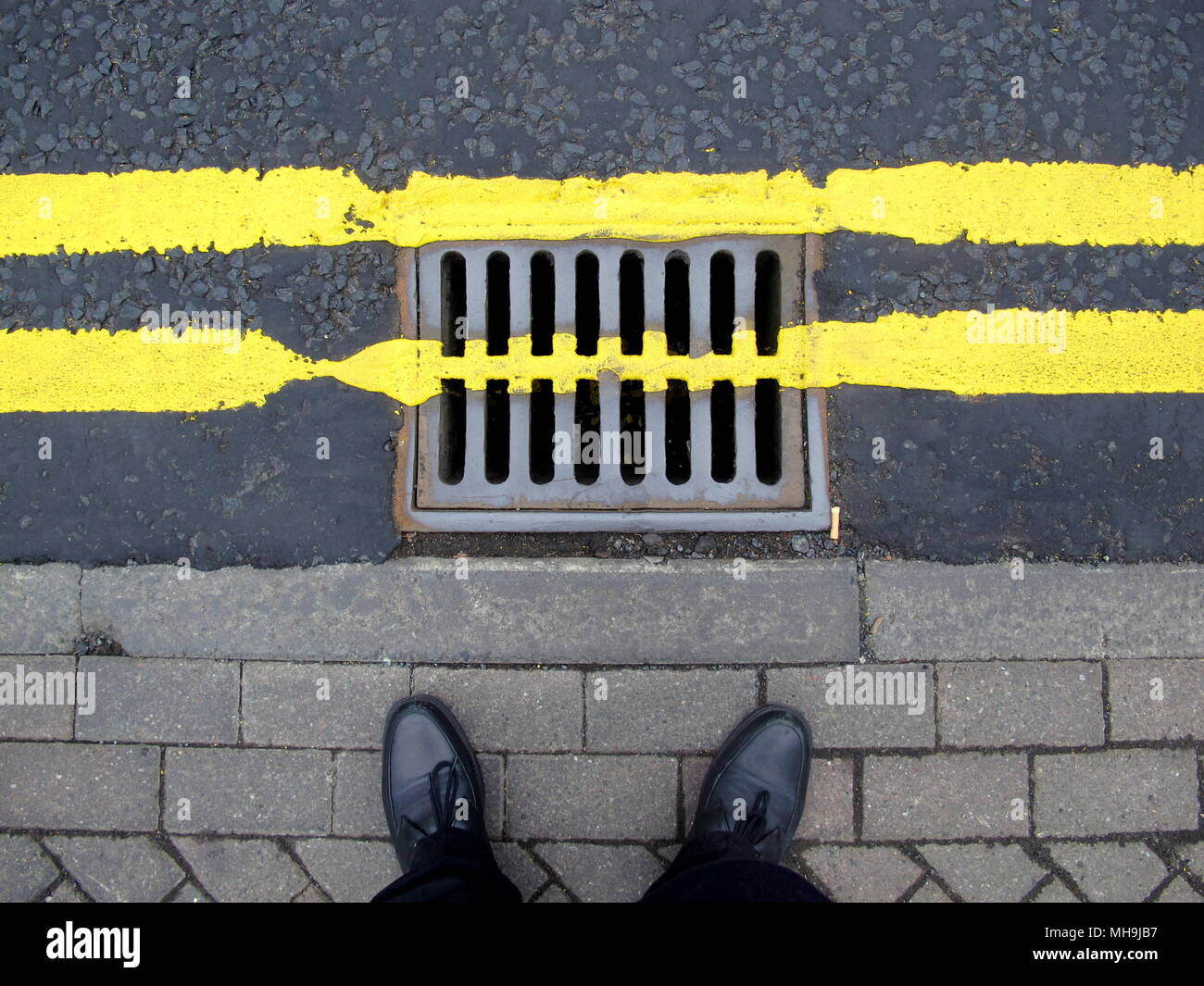 double yellow lines pavement sidewalk feet shoes with grating drain cover painted over road street Stock Photo