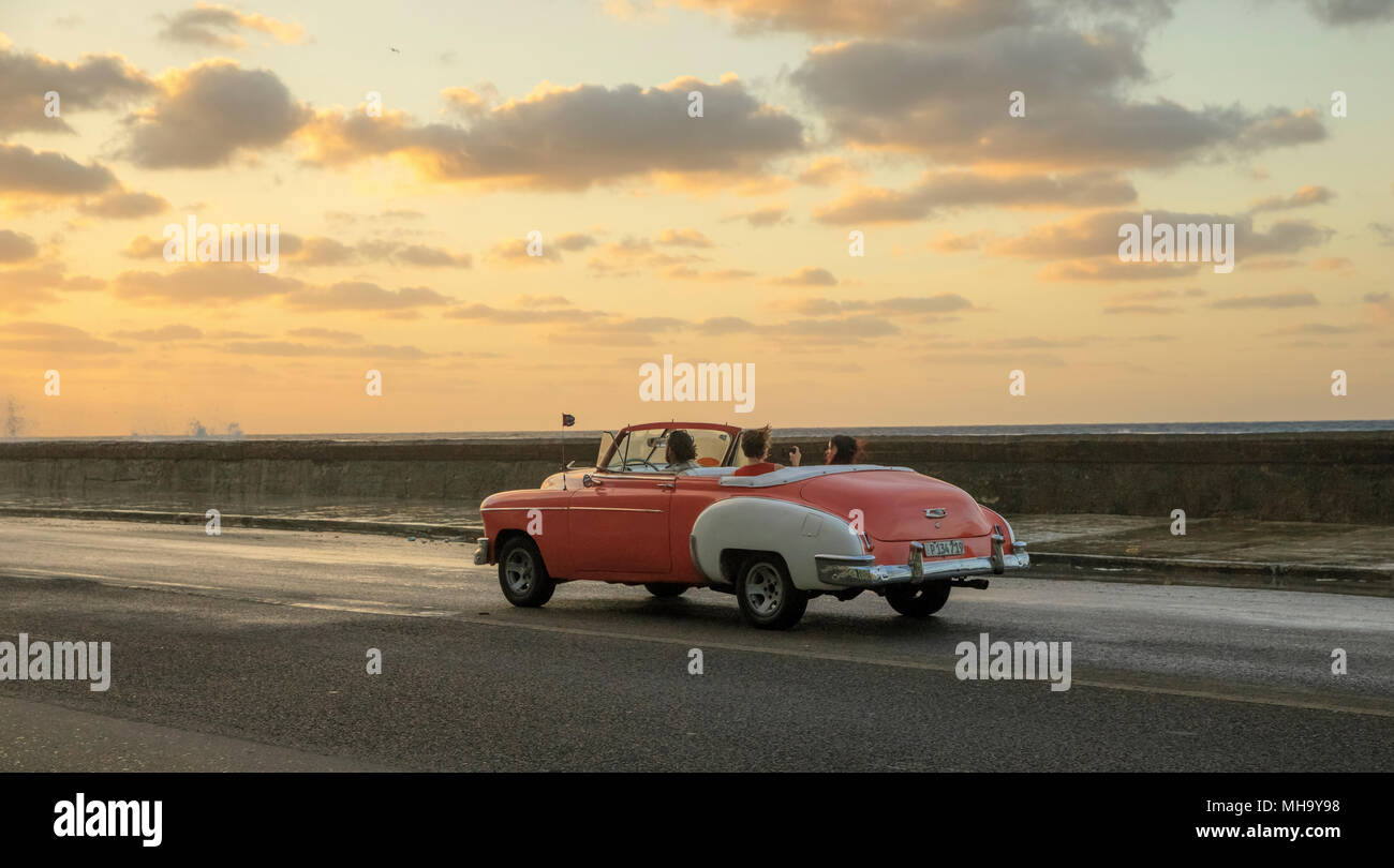 Waves crash as the sun sets as classic cars travel along the Malecon in Havana, Cuba. Stock Photo