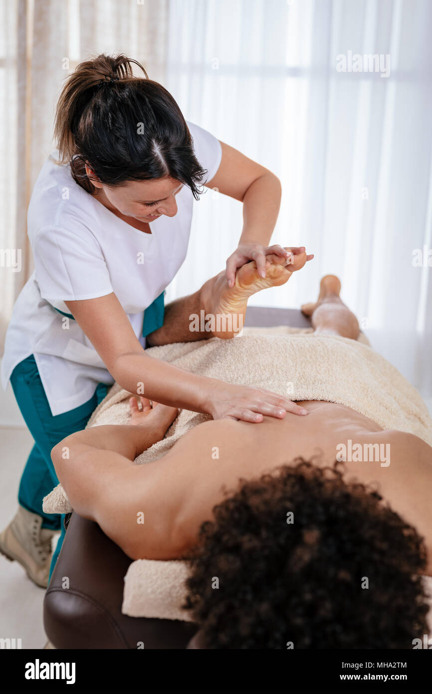 Handsome man lying down on massage table and getting healthy leg massage by young female therapist in the spa center. Stock Photo