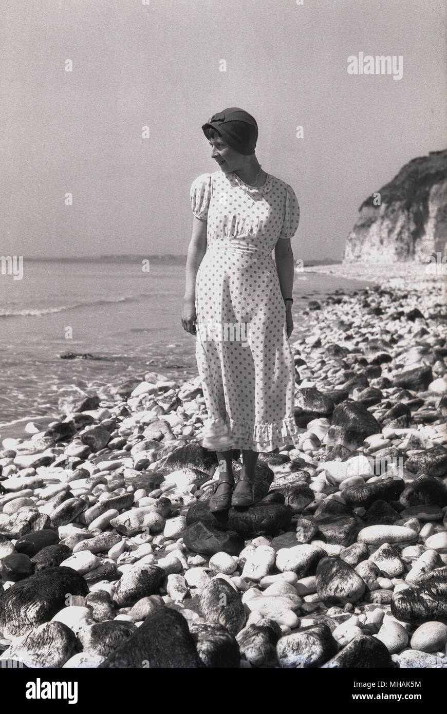 1930s, historical, lady standing on a pebbly beach wearing the latest fashions, a long polka-dot dress, shoes and hat, England, UK. Stock Photo