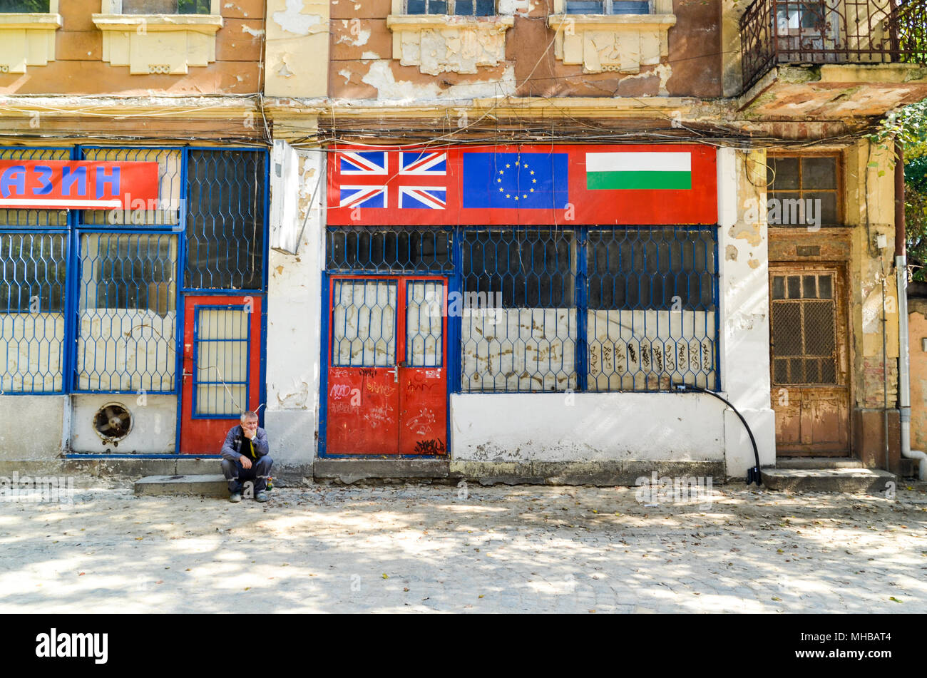 Old man sitting by a street, in front of a shop with the Bulgarian, UK and EU flags. Plovdiv, Bulgaria Stock Photo