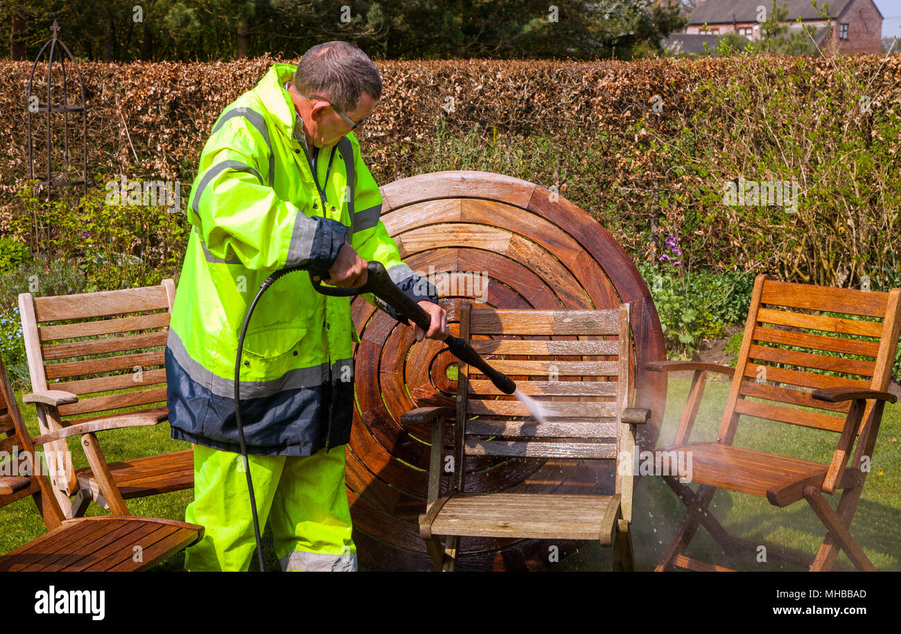 Man wearing high visibility clothing and workwear cleaning hardwood wooden garden furniture with a power / jet washer Stock Photo