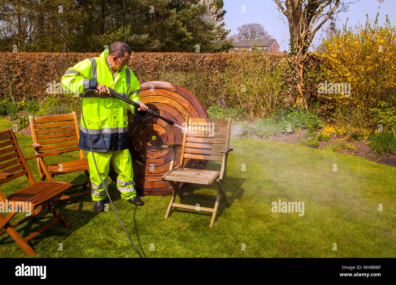 Man wearing high visibility clothing and workwear cleaning hardwood wooden garden furniture with a power / jet washer Stock Photo