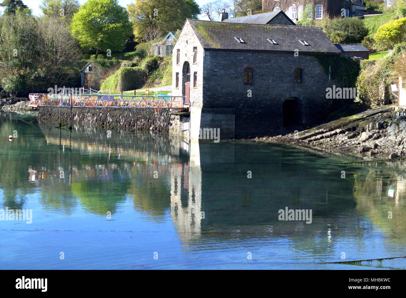 Stone built boat house reflected in the blue calm waters of castlehaven harbour, ireland at low tide. The harbour is a popular holiday destination Stock Photo