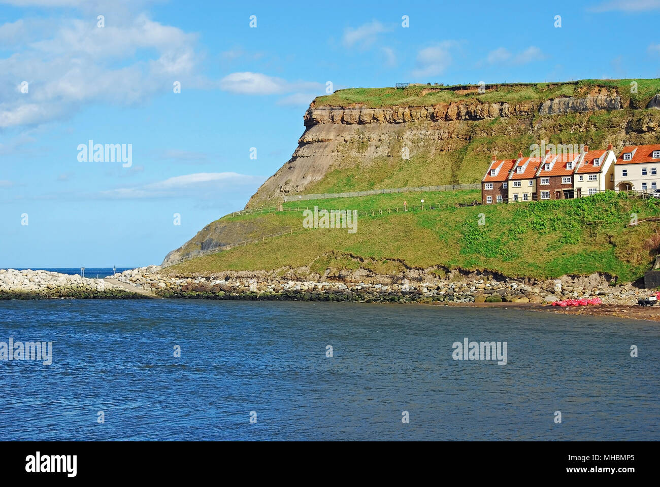 A grassy headland seen under a blue sky across the River Esk from Whitby town, with a row of red roofed houses situated on the headland Stock Photo