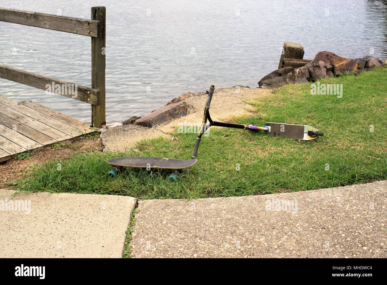 Skateboard and scooter lying on grass in park near water. Abandoned