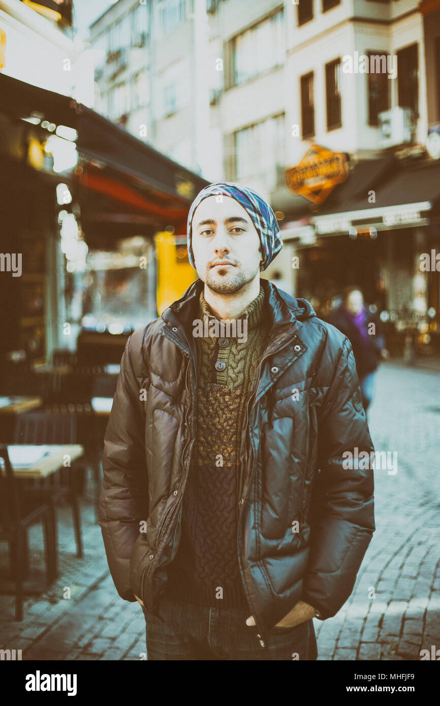 A young man in street with head scarf looking at camera with black jacket and blurred background Stock Photo