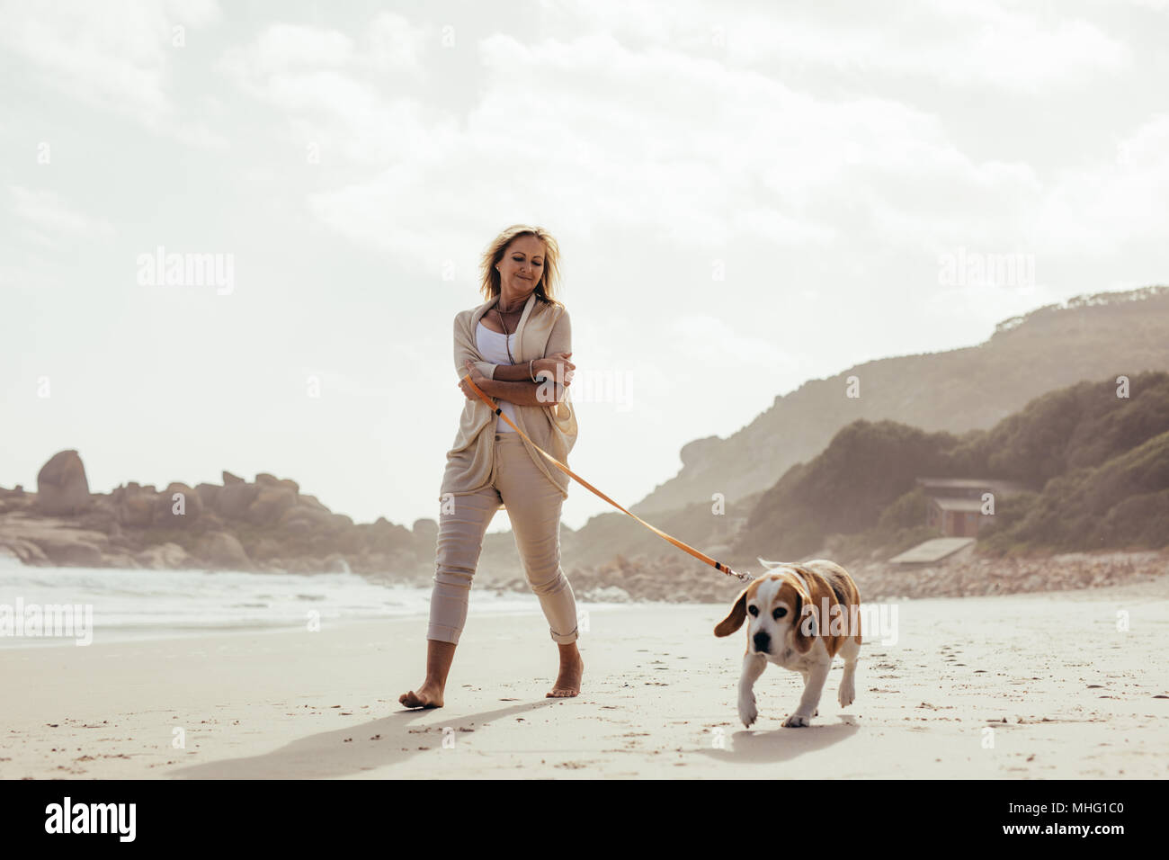 Full length shot of mature woman taking her dog on walk on the beach. Senior female on morning walk with her pet dog. Stock Photo