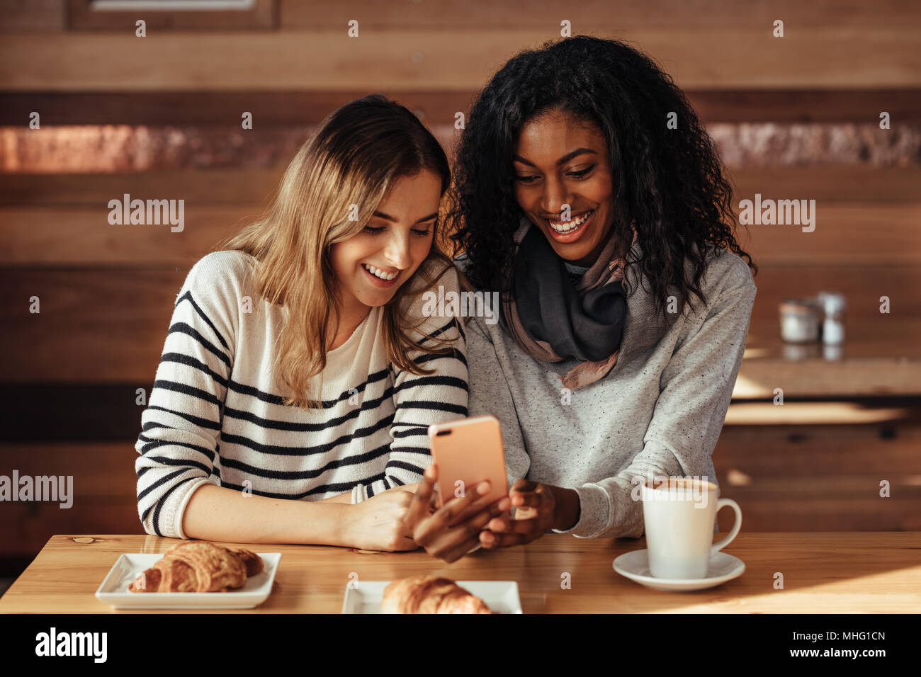 Two smiling women sitting in a restaurant looking at mobile phone. Friends sitting at a cafe with coffee and snacks on the table looking at mobile pho Stock Photo