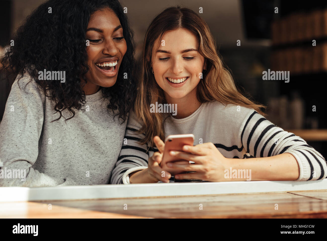 Two smiling women sitting in a restaurant looking at mobile phone and laughing. Friends sitting at a cafe smiling looking at a mobile phone. Stock Photo