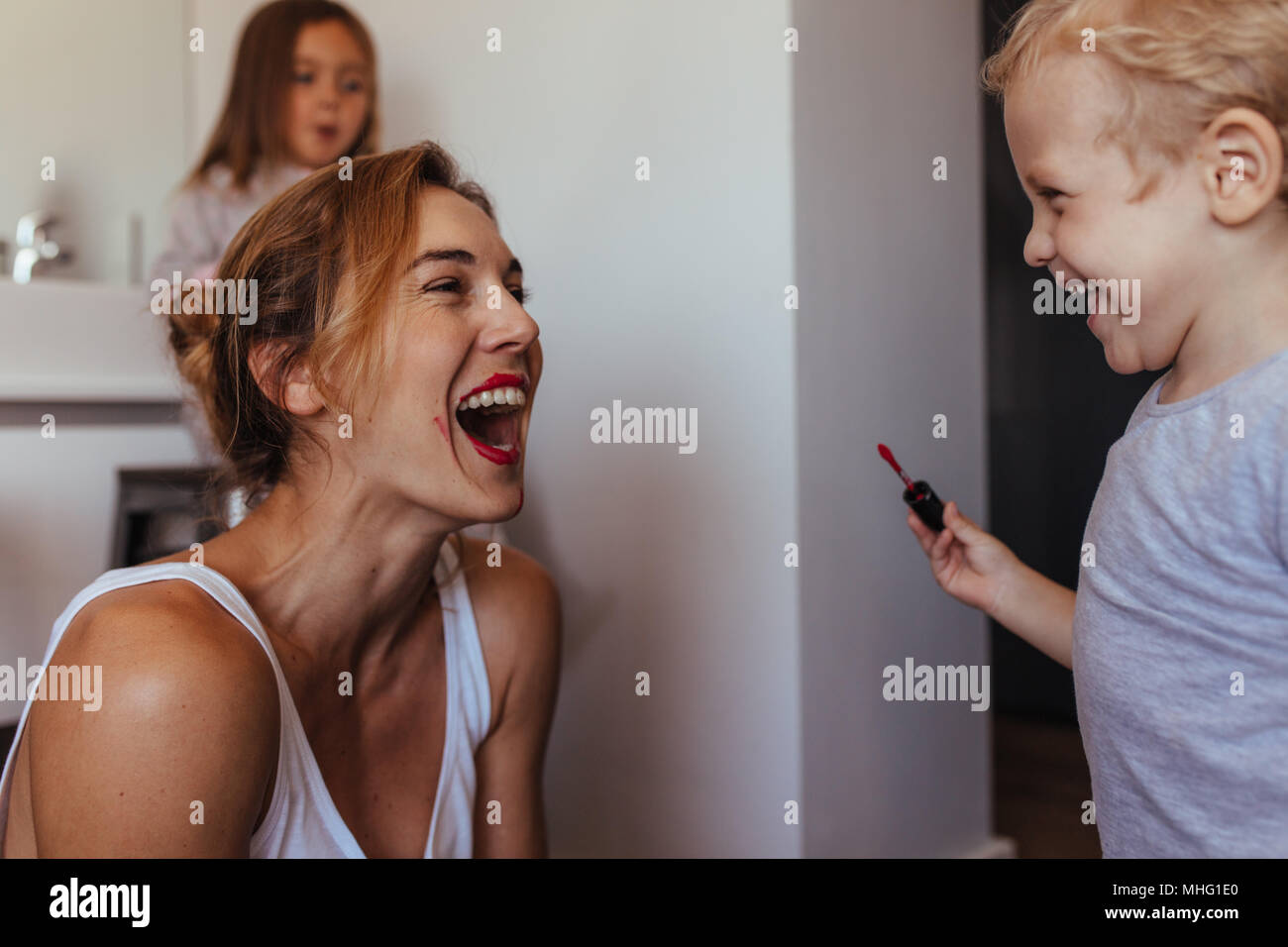 Little boy putting makeup on his mother's face and laughing with a girl sitting at the back in the bathroom. Mother and son playing with makeup indoor Stock Photo