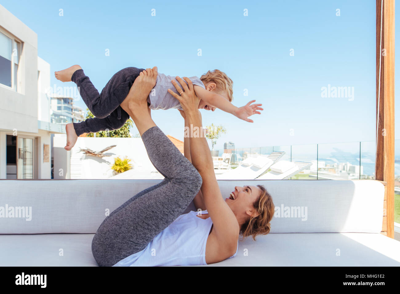 Side view of happy young mother playing with son on the sofa. Woman lying on couch and lifting a little boy with her legs. Stock Photo