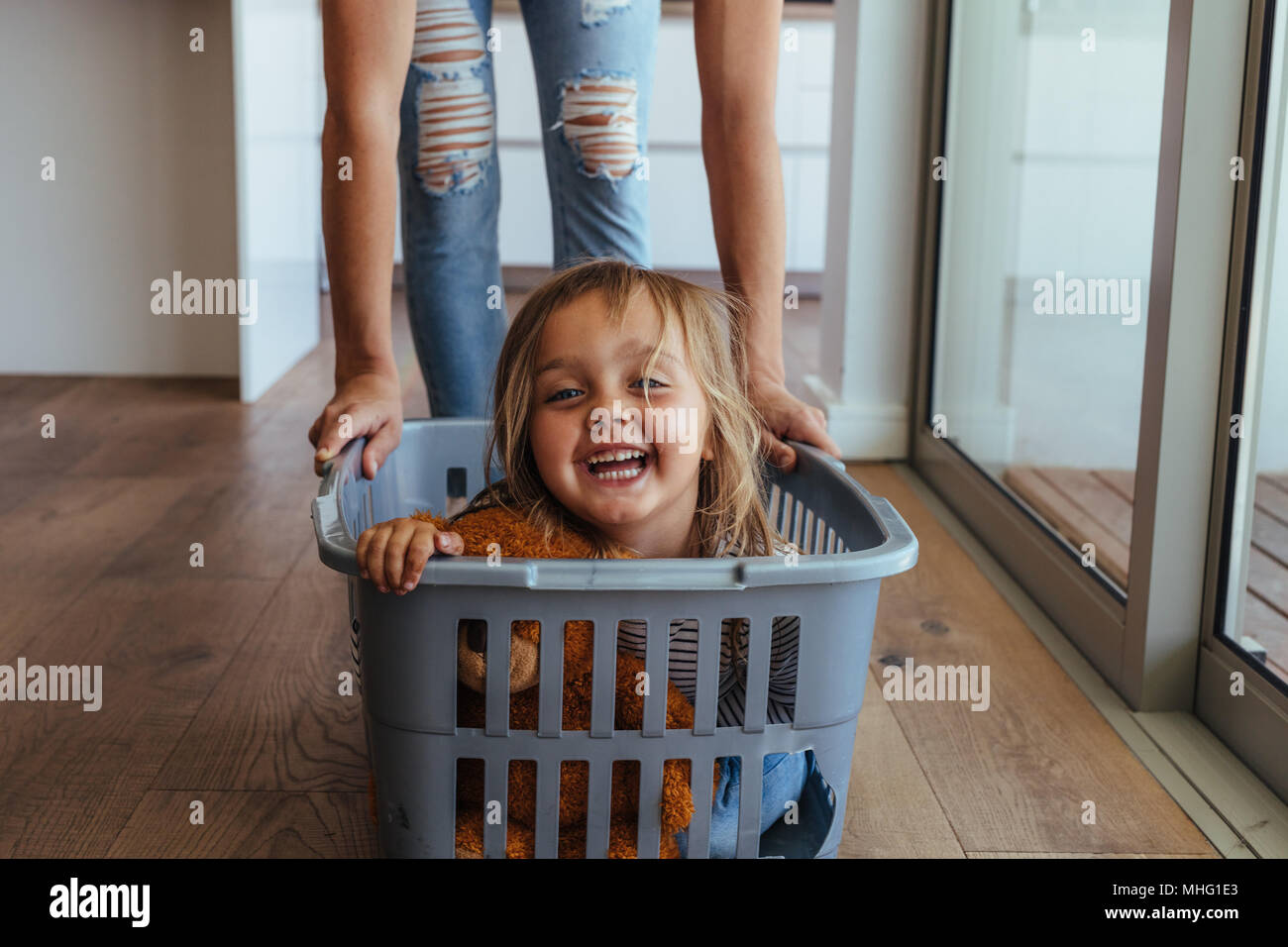 Beautiful little girl sitting in a washing basket being pushed by her mother. Cute little girl and her mother playing while doing laundry at home. Stock Photo