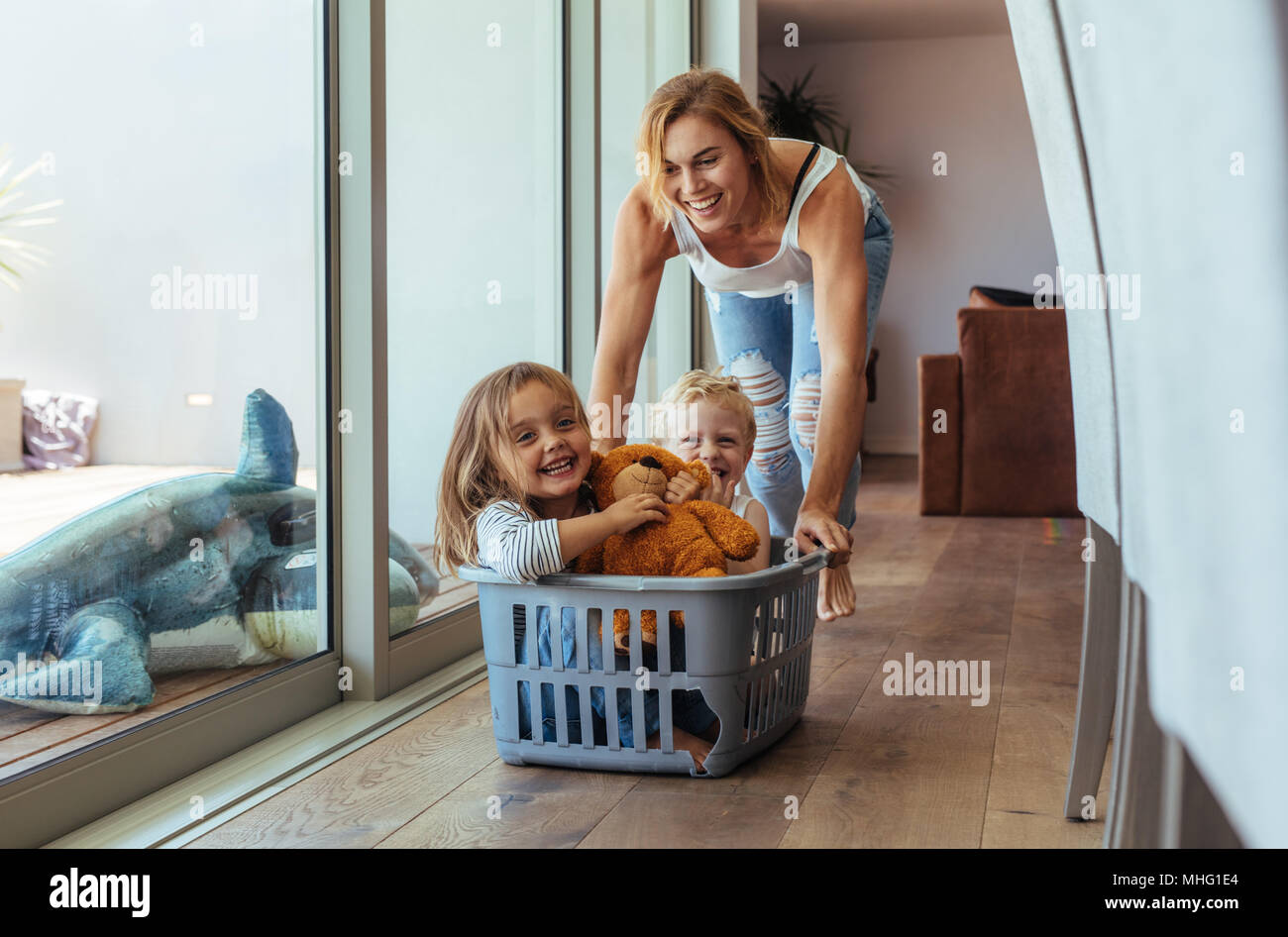 Happy young mother pushing children sitting in laundry basket. Mother and children playing at home. Stock Photo