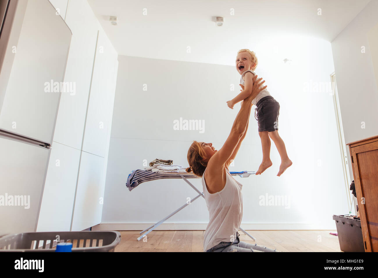 Smiling young woman holding her son in air. Mother and son playing in laundry room. Stock Photo