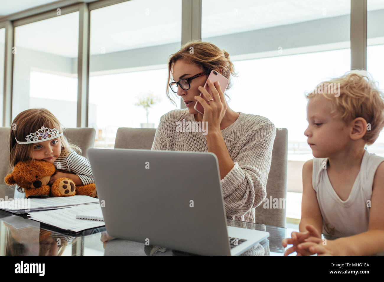 Young woman talking on mobile phone while sitting at tablet with laptop and her kids. Mother working from home with her children sitting by. Stock Photo