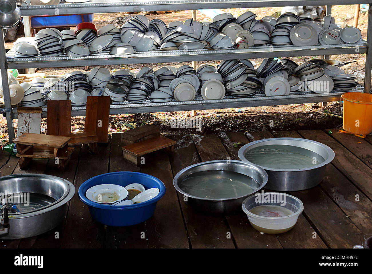 Plates and bowls are stacked on stainless steel shelves. There is a bowl of dishes put in basin. Basin with water. Small wooden chairs and yellow tank Stock Photo