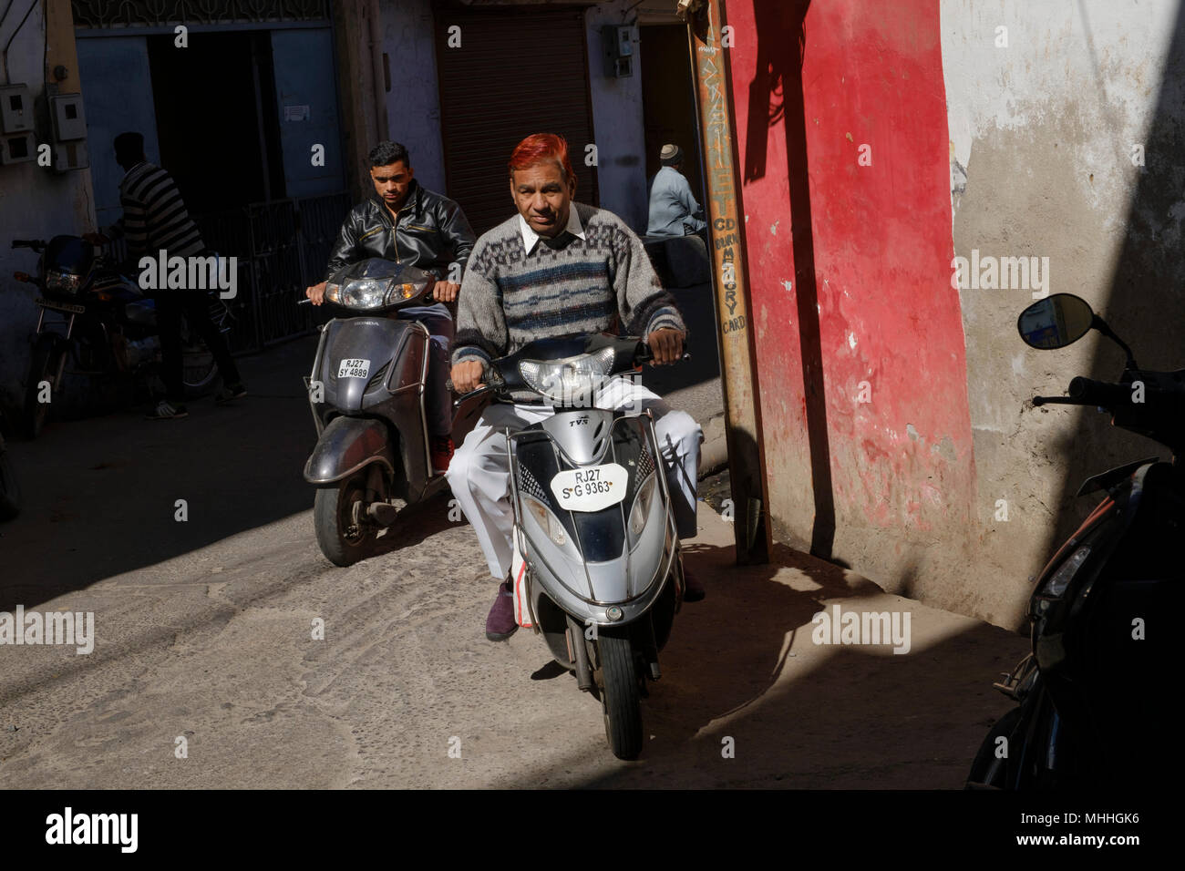 Man with red hair on a motorbike passing a red wall.. Udaipur, also known as the City of Lakes, The Venice of the East, is the historic capital of the kingdom of Mewar, Rajasthan. Stock Photo