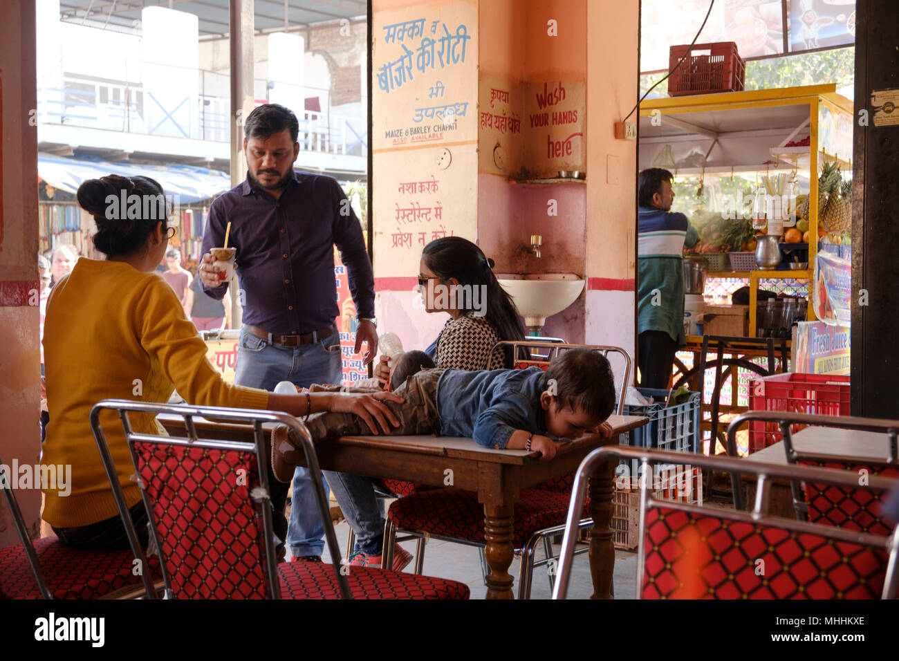 India - Pushkar - Child lies on a table in cafe as man brings a drink to the table. Stock Photo