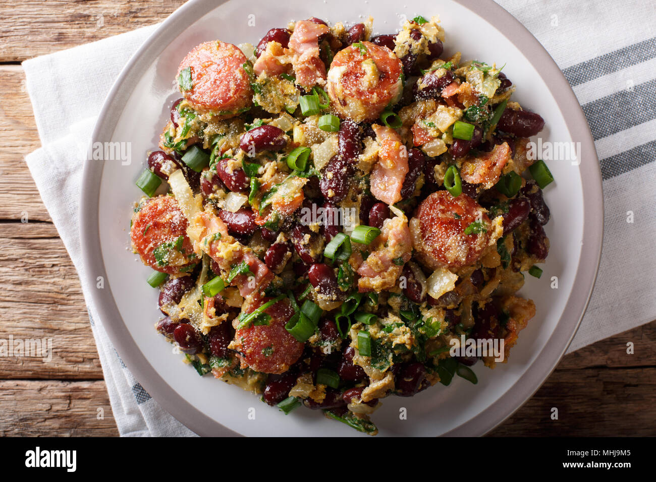 Brazilian beans with greens, sausages, bacon and eggs close-up on a plate on a table. Horizontal top view from above Stock Photo