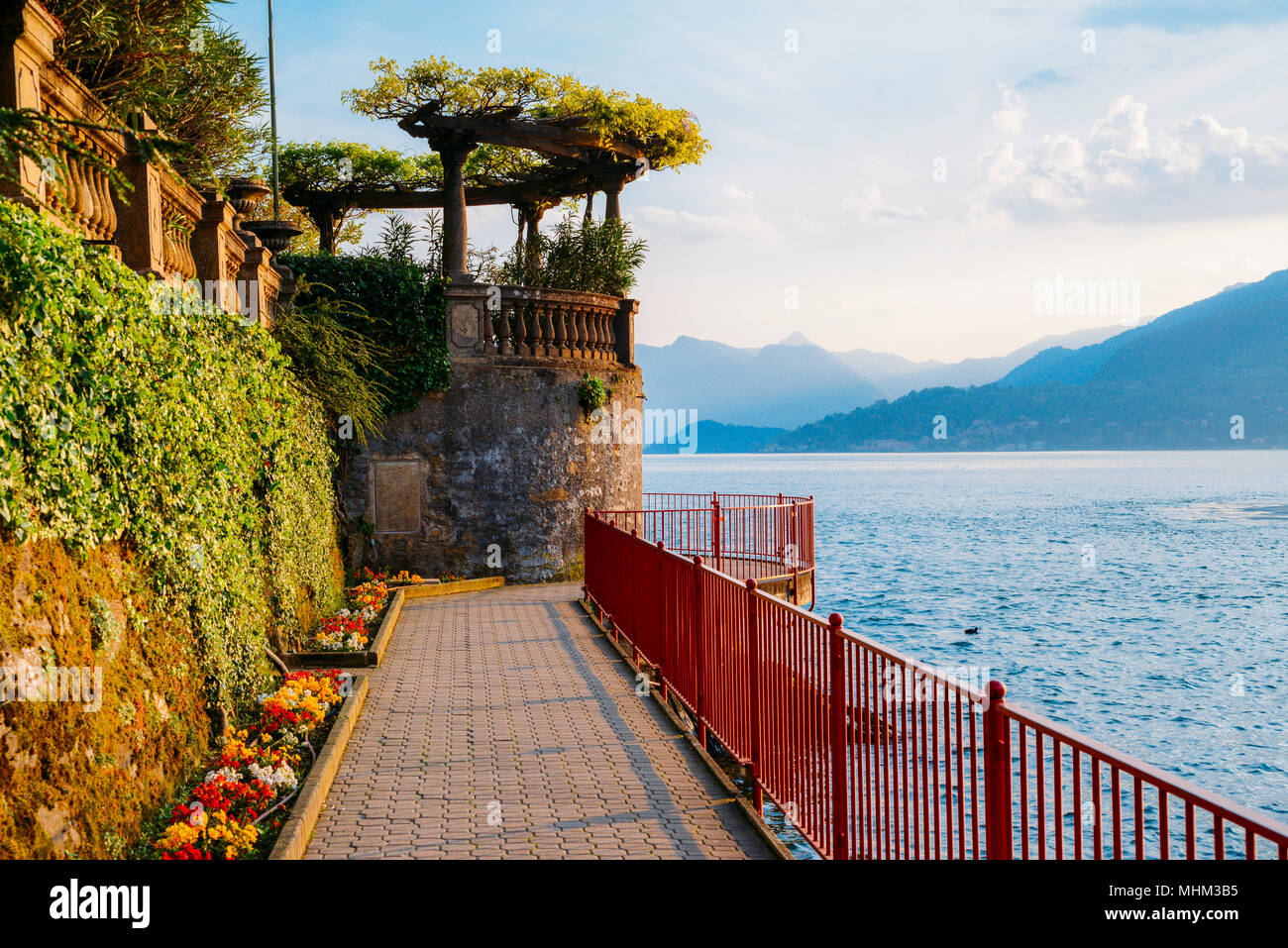 Red garden arch on coastline leading towards the beautiful and historic city of Varenna on the edge of Lake Como in the northern Italian region of Lombardy. Stock Photo