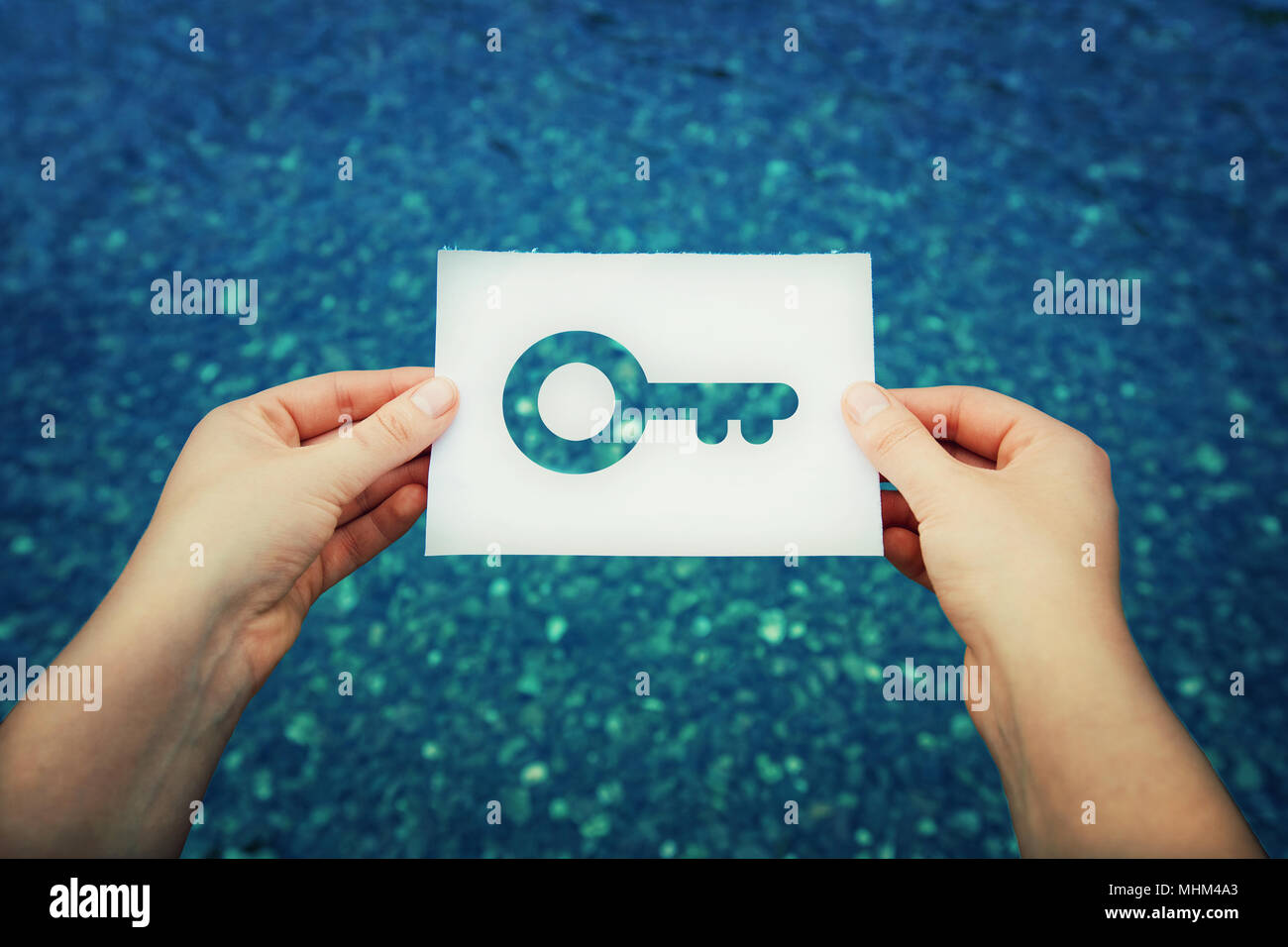 Close up of woman hands holding a white paper sheet with a key icon insideon a blue sea water background. Unlock success and feel free. Stock Photo